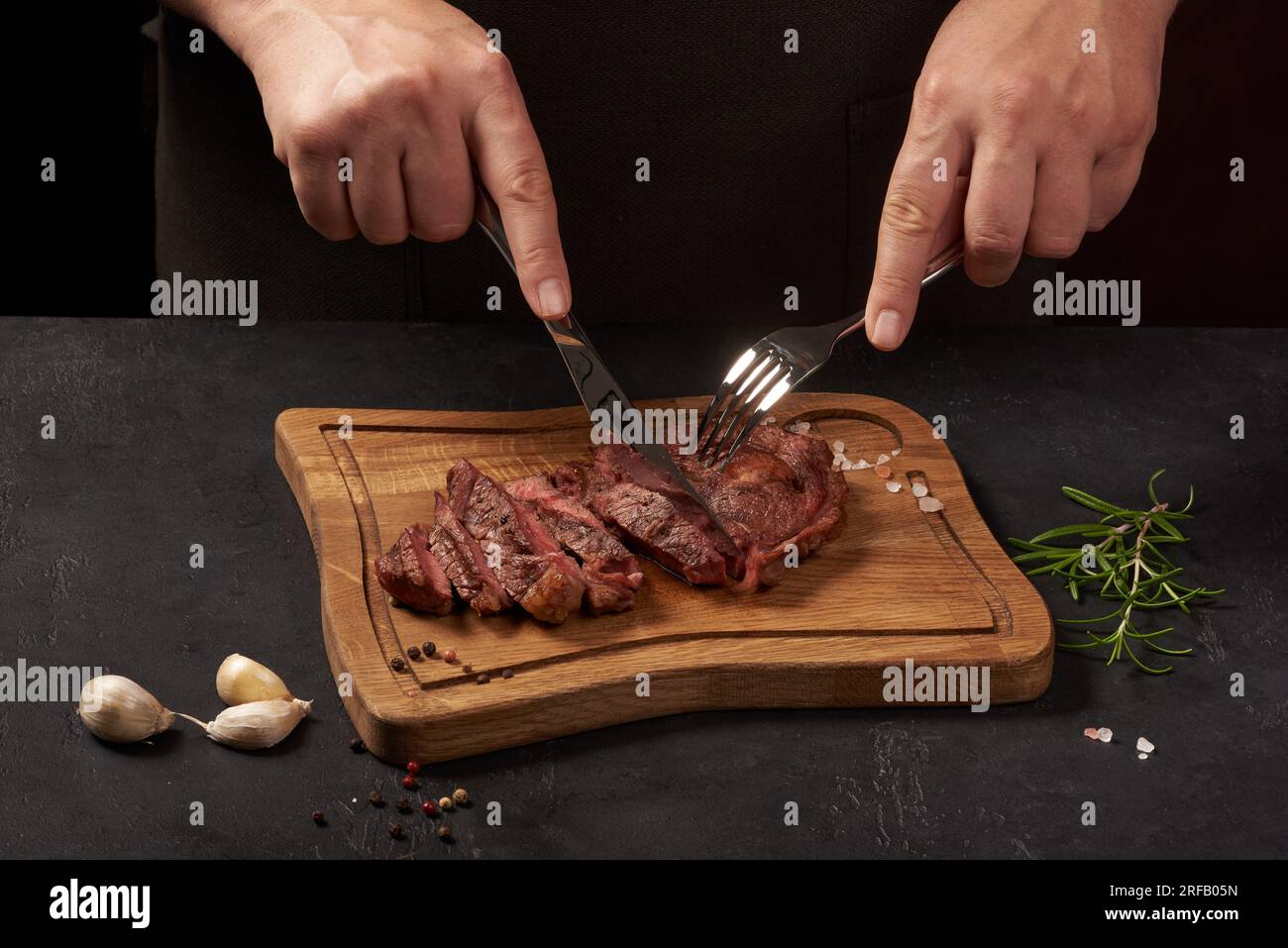 Gegrilltes Rindersteak, das in Scheiben auf einem Holzschreibtisch mit schwarzem Hintergrund geschnitten wird, mit Gabel und Messer. Der Mann schneidet Fleisch. Stockfoto