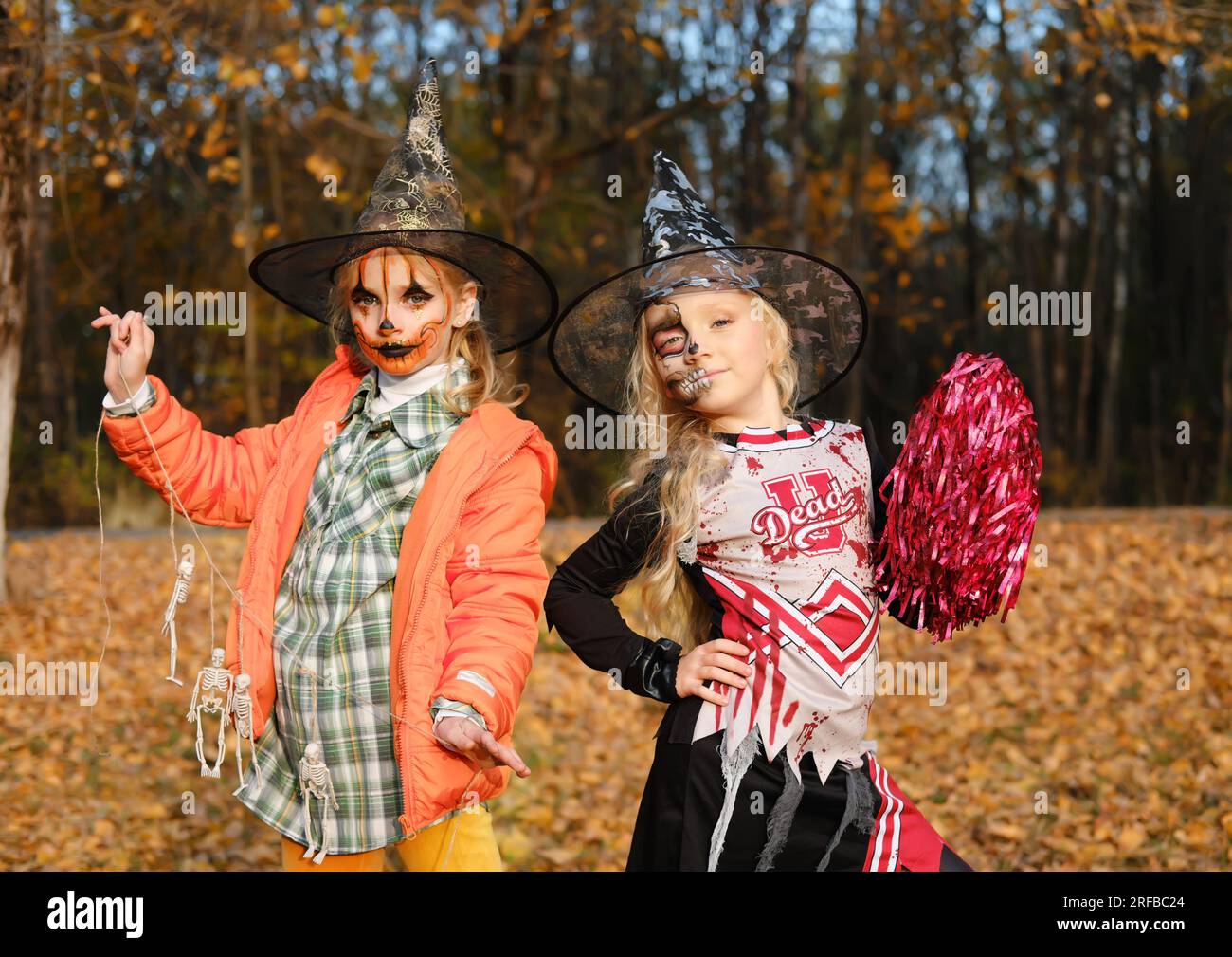 Ein Mädchen in einem Cheerleader-Kostüm mit halbgesichtlichem Skelett-Make-up und ein Mädchen mit Kürbis-Make-up in Hüten posieren und in die Kamera schauen. Horizontales Foto Stockfoto