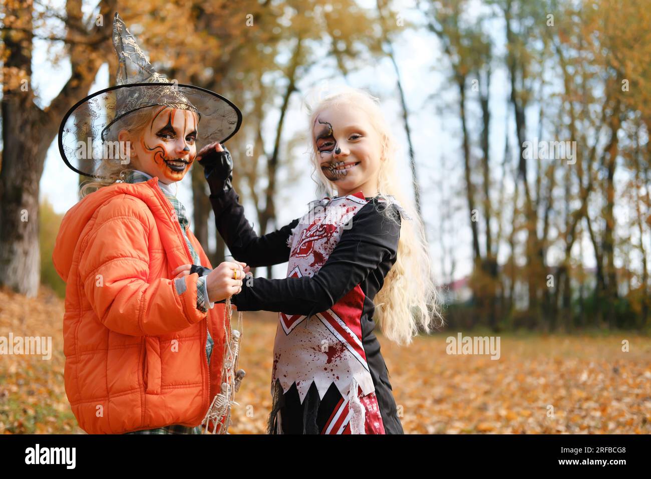 Ein Mädchen, das als Cheerleaderin verkleidet ist und halb-Gesicht-Make-up in Form eines Skeletts hat, passt den Hut eines Mädchens in Form eines Kürbels an. Lächelnde Mädchen Stockfoto