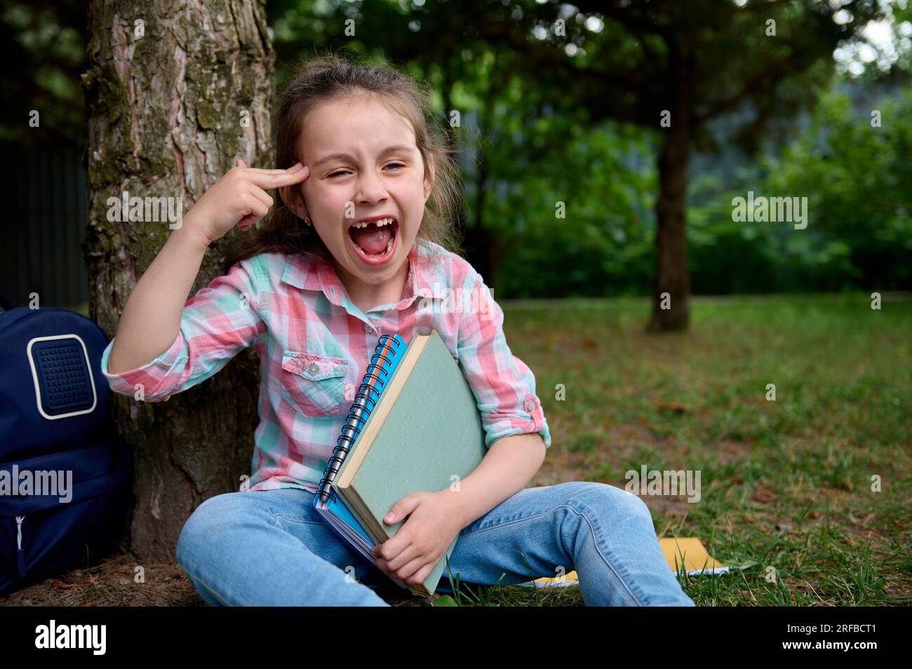 Ein bösartiges kleines Mädchen, das Schulbedarf in der Hand hat, sitzt unter einem Baum im Park, Gesten, zeigt mit dem Finger auf ihren Tempel, grimpert, macht Gesicht Stockfoto