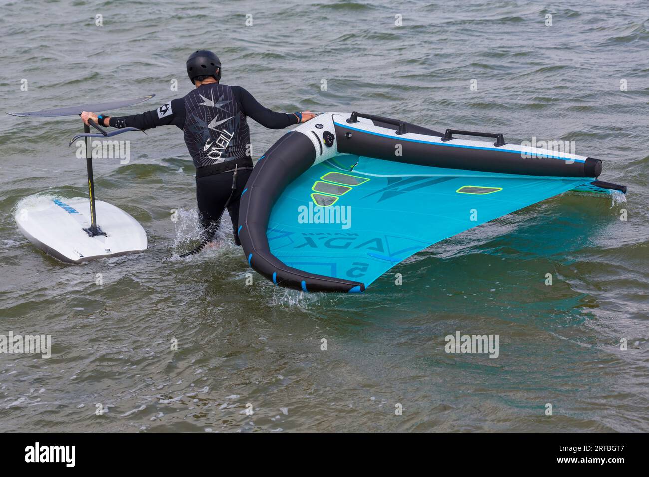 Ein Mann trägt einen O'Neill Neoprenanzug und trägt Flügelfolien ins Meer, bereit für Flügelfolien im Juli in Sandbanks, Poole, Dorset, Großbritannien Stockfoto