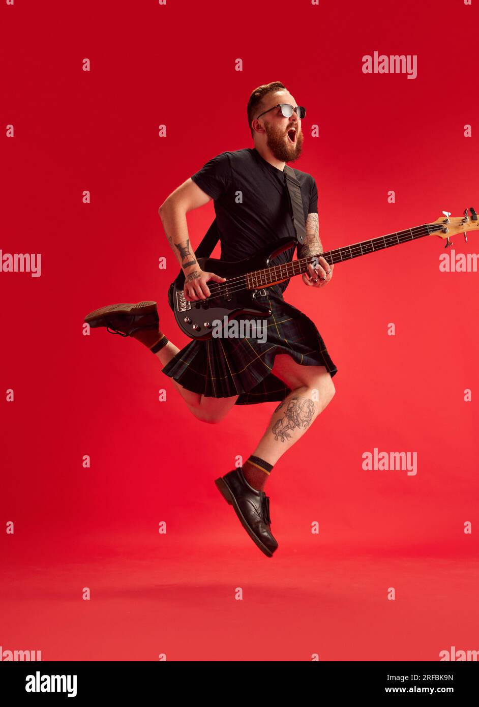 Bärtiger Mann in Sonnenbrille und Rock, Kilt spielt Gitarre vor rotem Studiohintergrund. Emotionaler Musiker Stockfoto