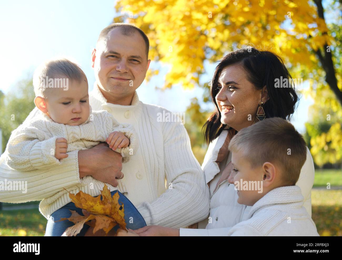 Eine glückliche vierköpfige Familie verbringt Zeit im Herbstpark. Mutter und ältester Sohn schauen auf das jüngste Kind, Vater lächelt und schaut in die Kamera. Horizont Stockfoto