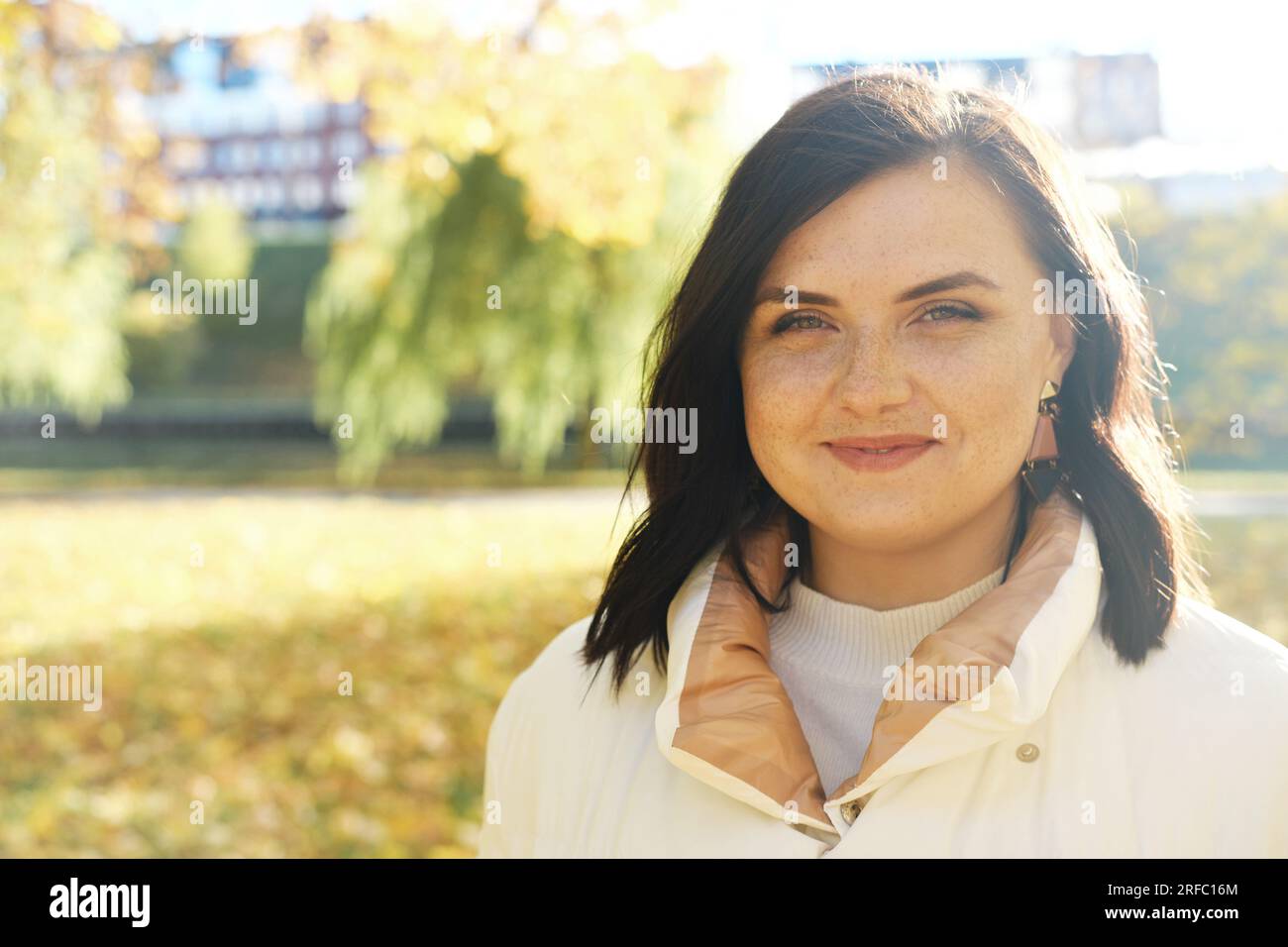 Schöne braune Frau mit Sommersprossen im Gesicht lächelt und schaut in die Kamera. Junge Frau, die bei sonnigem Wetter im Herbstpark posiert. Horizontales Foto Stockfoto