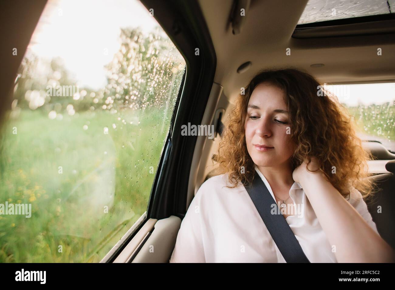 Fröhliche, positive, lockige junge Frau in Freizeitkleidung, die auf dem Rücksitz sitzt, den Sicherheitsgurt anschnallt und wegschaut Stockfoto