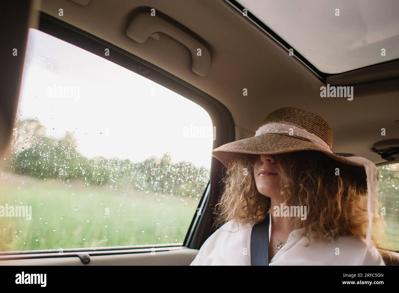 Fröhliche, positive, lockige junge Frau in Freizeitkleidung mit Hut sitzt auf dem Rücksitz des Autos mit angelegtem Sicherheitsgurt Stockfoto