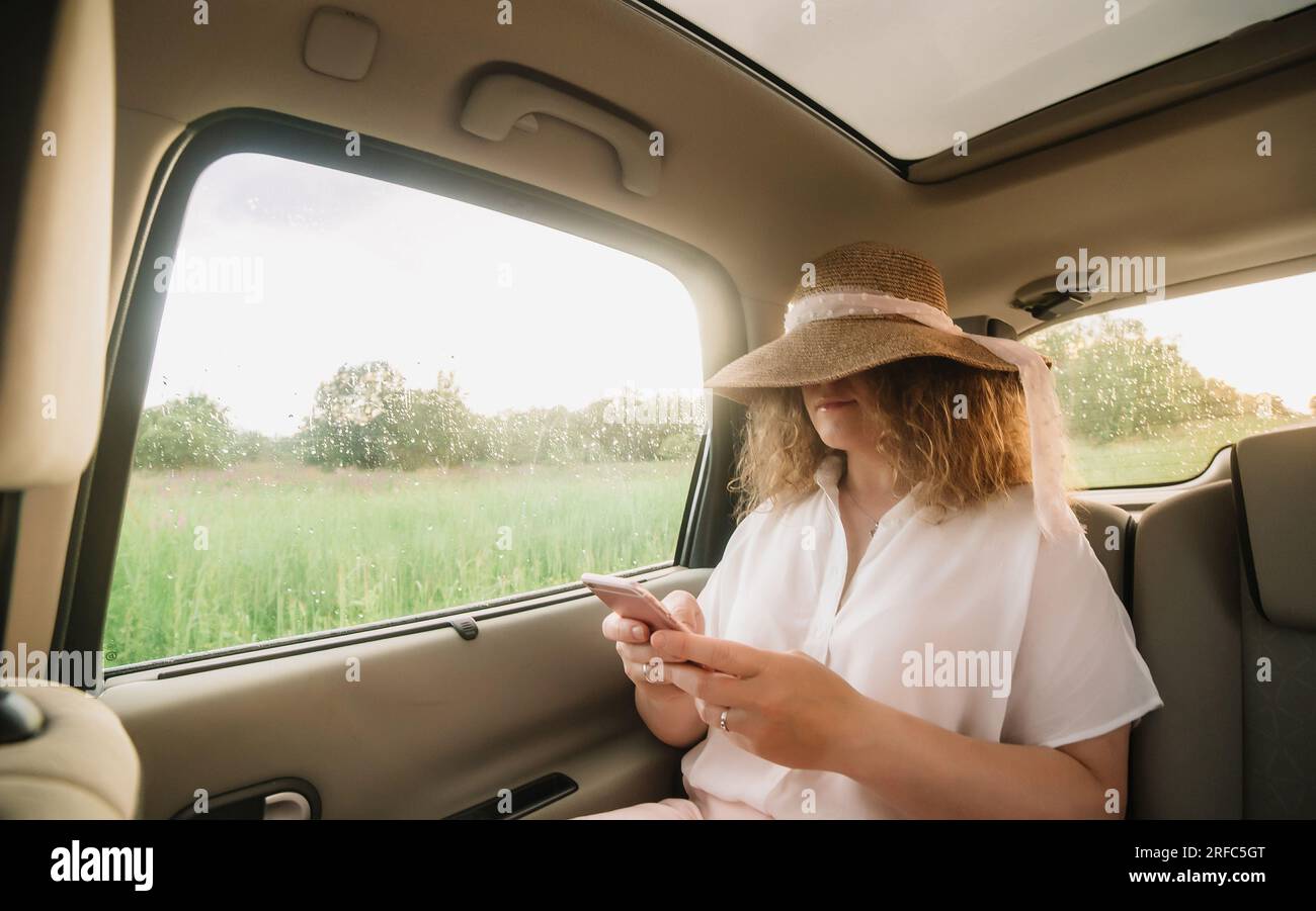 Fröhliche, positive, lockige junge Frau in Freizeitkleidung mit Hut, sitzt auf dem Rücksitz des Autos mit angelegtem Sicherheitsgurt und telefoniert Stockfoto