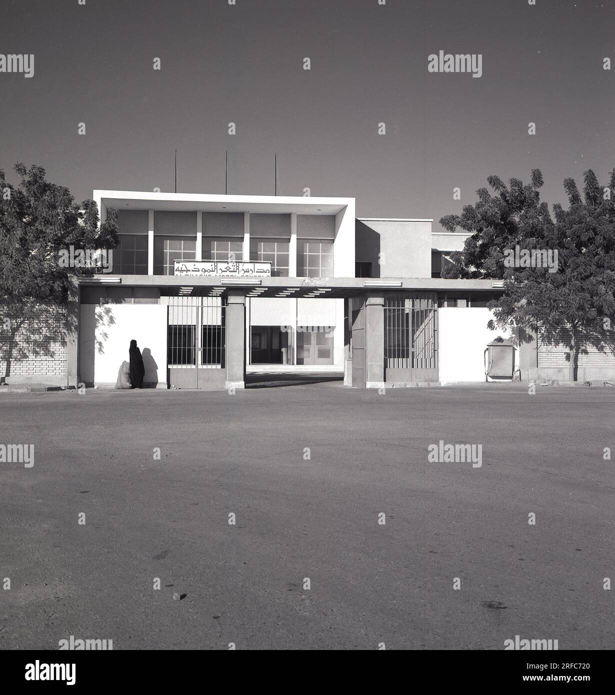 1960er Jahre, historische, arabische Frau, von Kopf bis Fuß mit einem langen schwarzen Gewand bedeckt, bekannt als arabischer Mantel oder Abaya, mit Sack, der vor dem Eingang zu einem modernen Gebäude steht, möglicherweise einer Bildungsschule, Saudi Arabien. Stockfoto