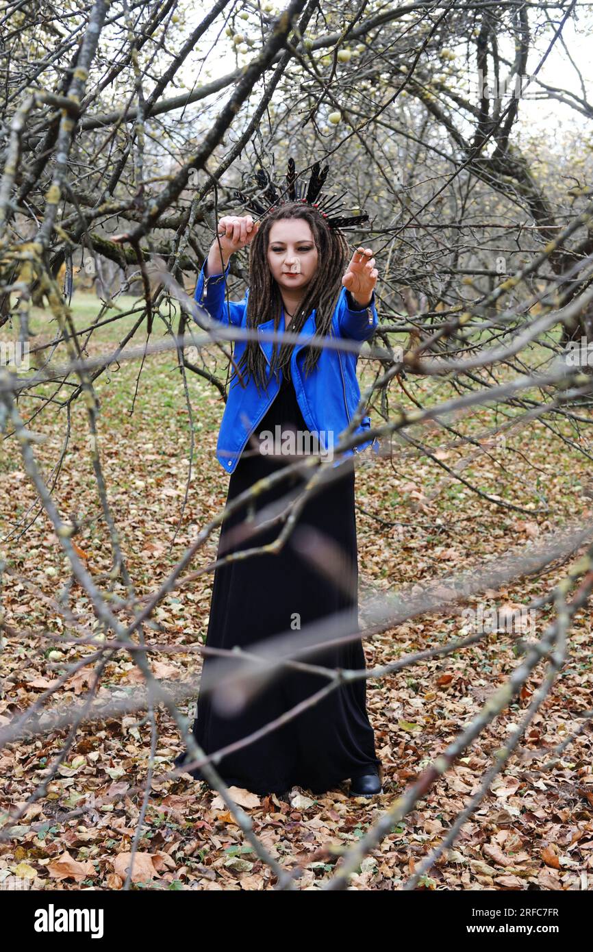 Porträt einer Frau in einer blauen Lederjacke mit Dreadlocks und einem Kopfband mit Federn. Eine Frau, die im Herbstpark posiert und in die Kamera schaut. Vertikal Stockfoto