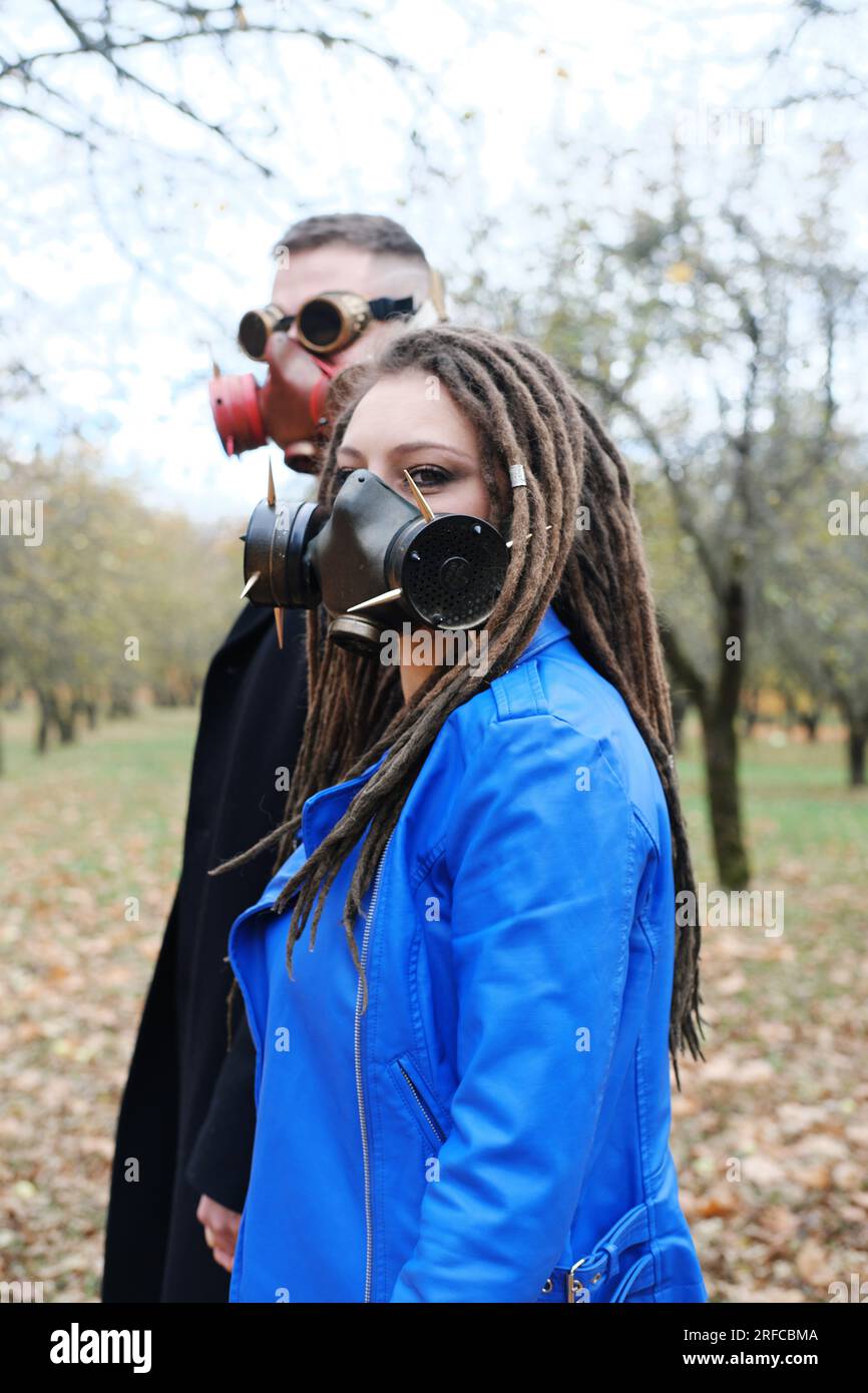Eine Frau mit Dreadlocks und einer Gasmaske und ein Mann mit einer Steampunk-Brille und einer Gasmaske Pose in einem Herbstpark. Ökologisches Katastrophenkonzept. Vertikal p Stockfoto