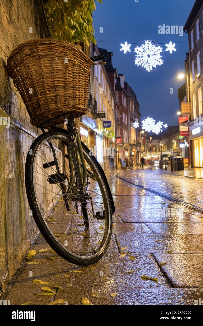 Das Fahrrad ruht an der Wand, auf der Sliver Street Cambridge, nasser Bürgersteig mit Weihnachtslichtern im Hintergrund. Stockfoto