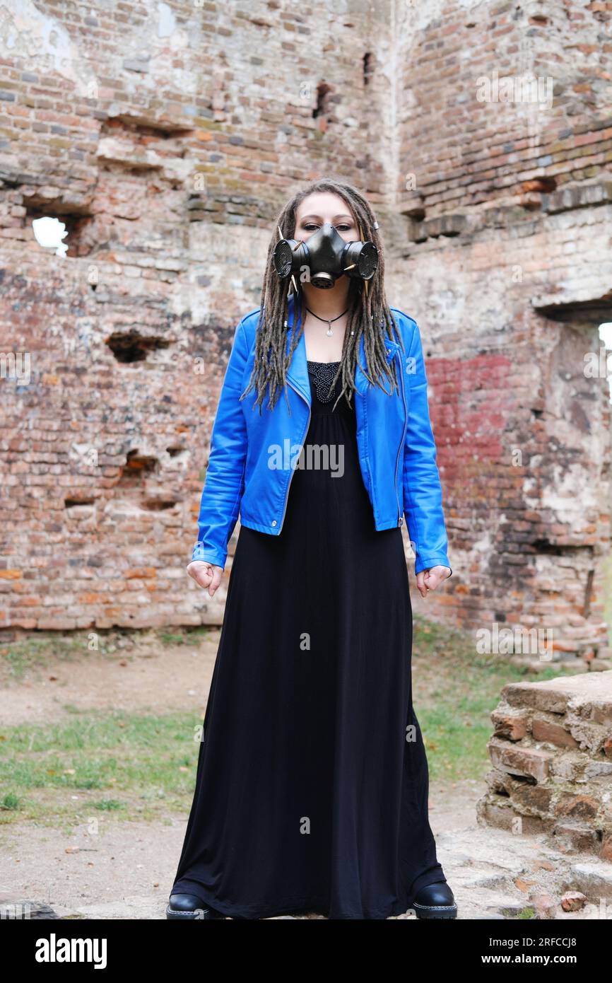 Frau trägt Gasmaske mit Stacheln in blauer Jacke und schwarzem Kleid, die in einem zerstörten Gebäude steht. Vertikales Foto Stockfoto