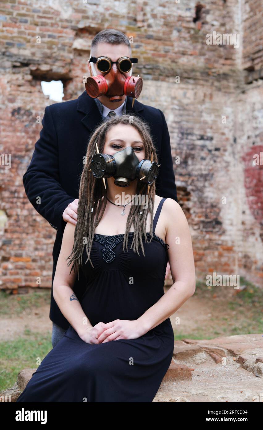 Ein junger Mann mit Steampunk-Schutzbrille, eine Gasmaske und eine Frau mit Dreadlocks und einer Gasmaske posieren vor dem Hintergrund einer zerstörten Ziegelwand. Vertica Stockfoto