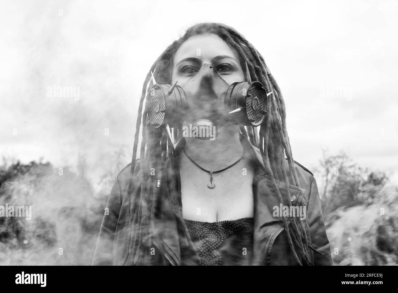 Porträt einer Frau in einer Jacke und Dreadlocks in einer Gasmaske mit Stacheln. Eine Frau, die in Rauch steht. Schwarzweißfoto. Horizontales Foto Stockfoto