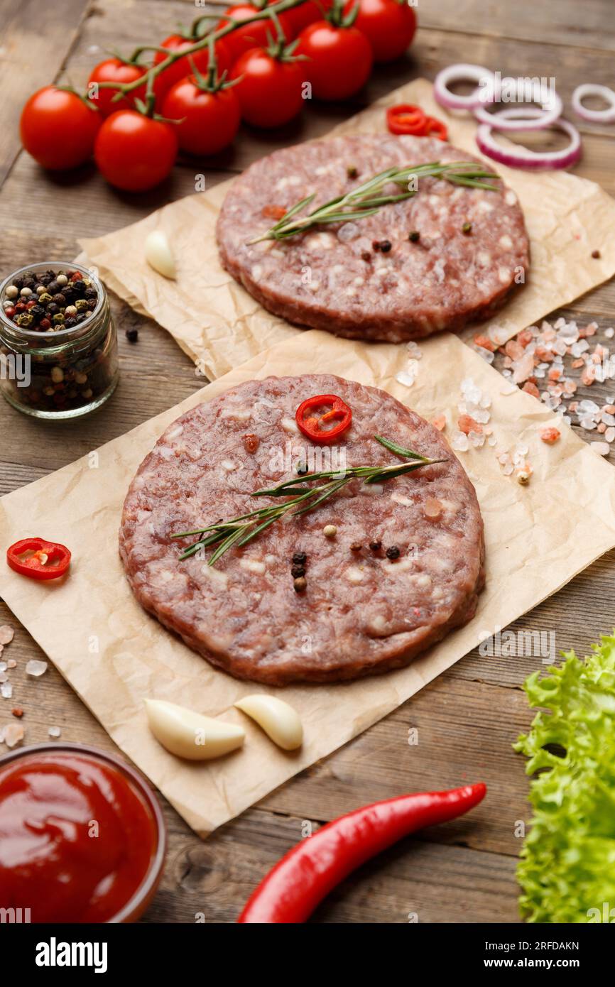 Rohe Burgerpasteten mit Gewürzen und Rosmarin auf Pergament, Sauce, frisches Gemüse, Hamburger-Zutaten auf Holzhintergrund. Stockfoto