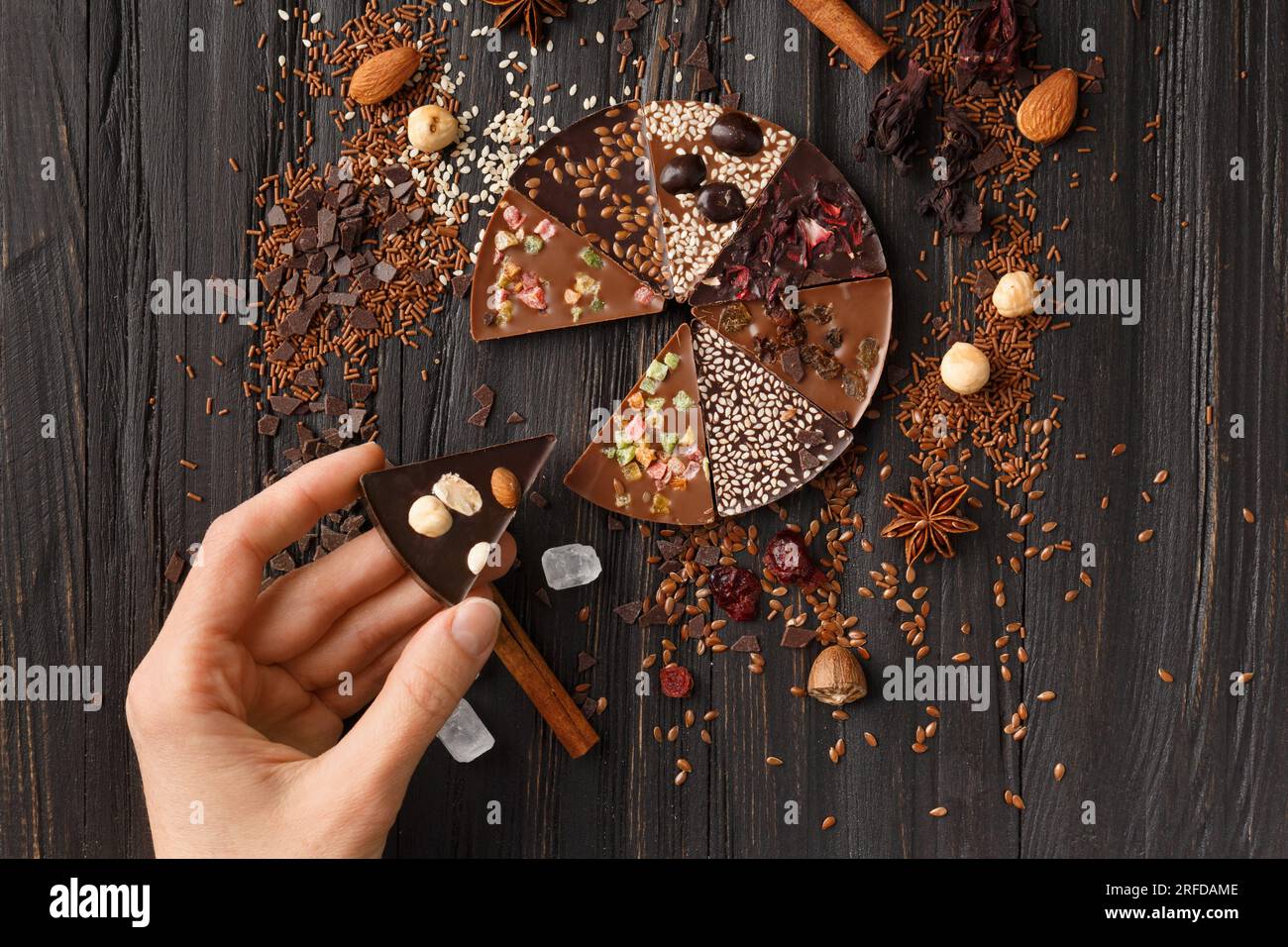 Schokoladenpizza mit Nüssen, trockenen Beeren, Sesamsamen und Zutaten auf dunklem Holzhintergrund von oben. Die Hand hält ein Stück süße Schokolade Stockfoto
