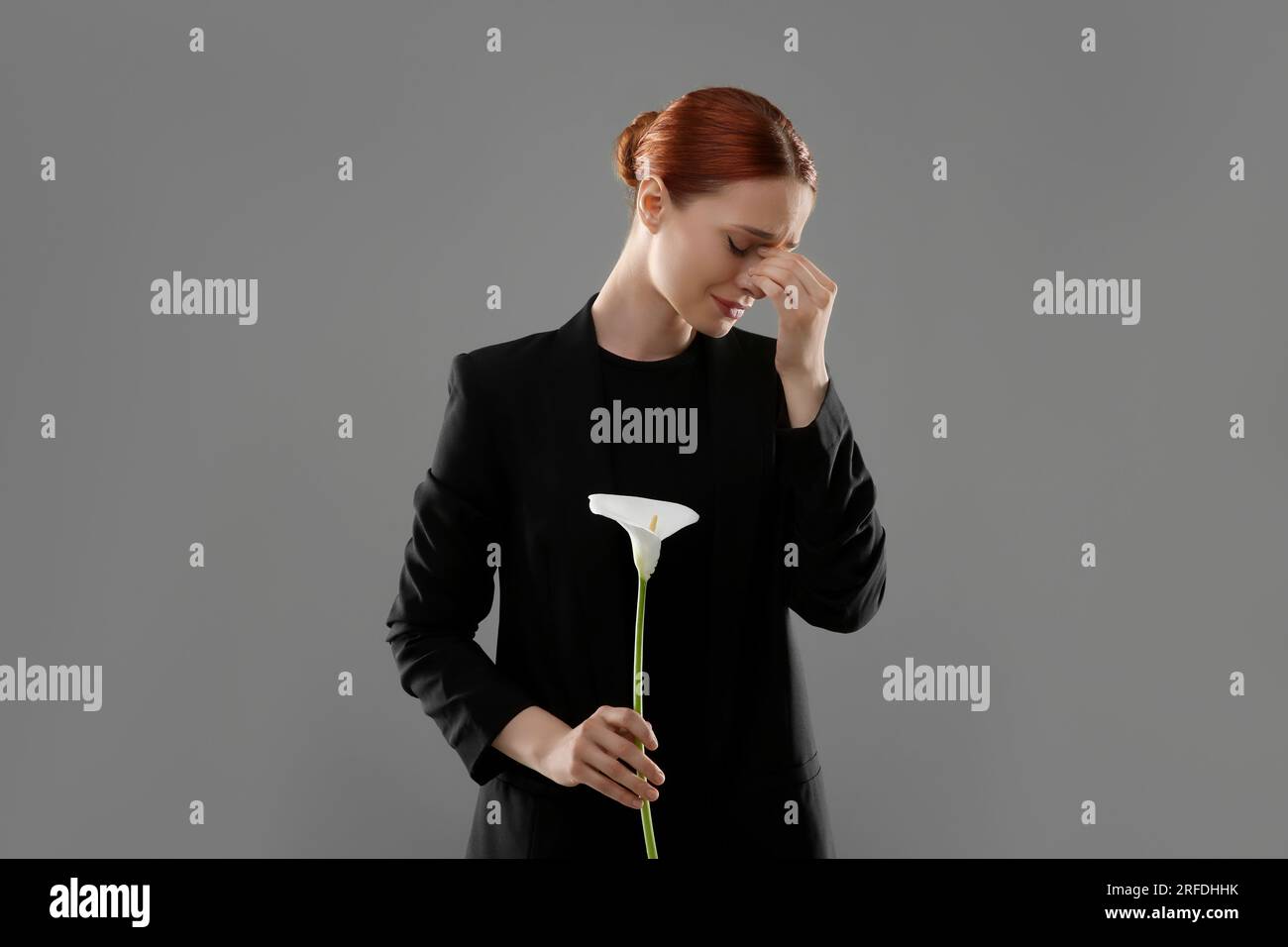 Traurige Frau mit Calla-Lilienblüten, die auf grauem Hintergrund trauert. Beerdigungszeremonie Stockfoto