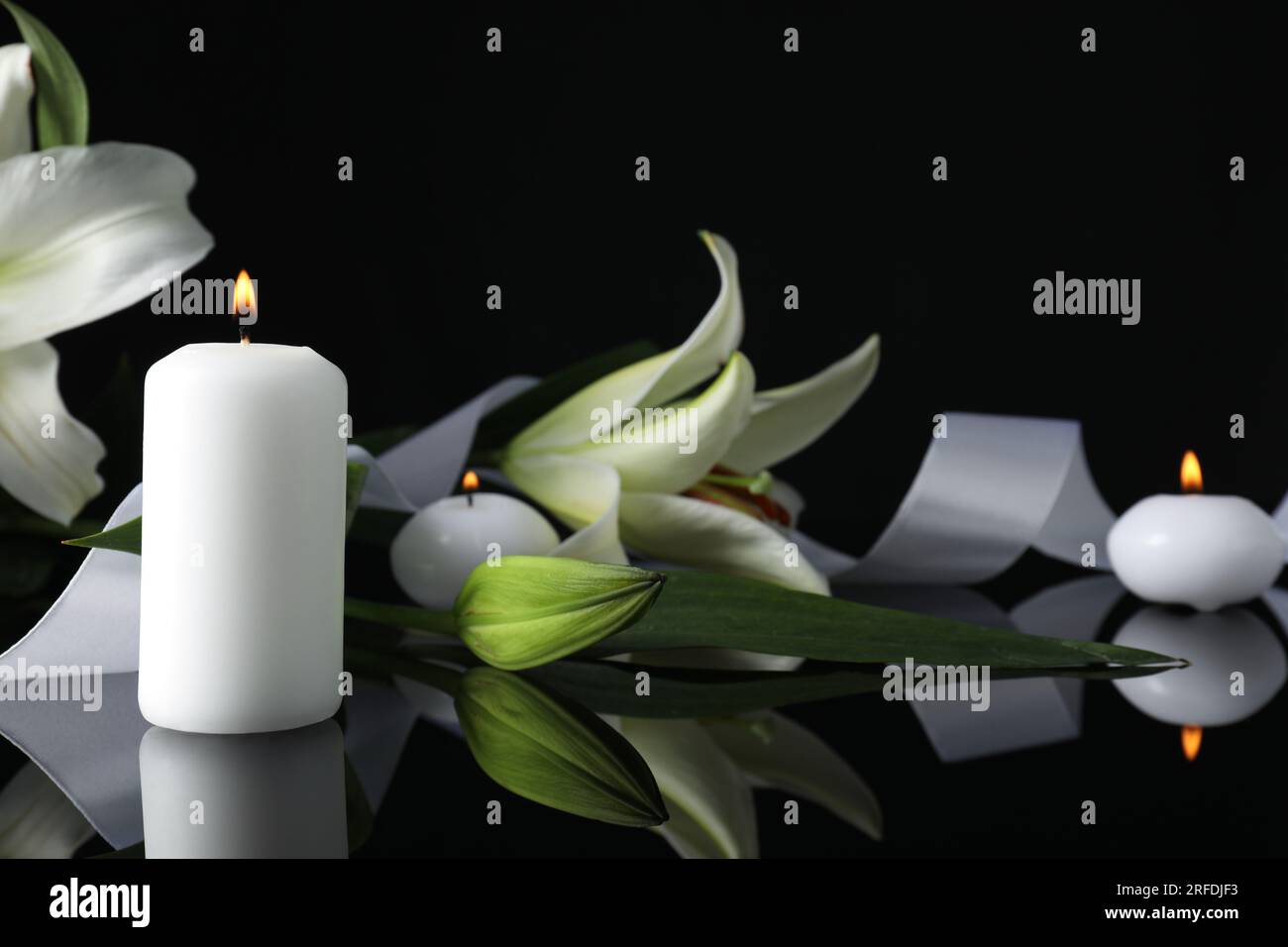 Weiße Lilien und brennende Kerzen auf schwarzer Spiegeloberfläche bei Dunkelheit, Nahaufnahme mit Platz für Text. Begräbnissymbole Stockfoto