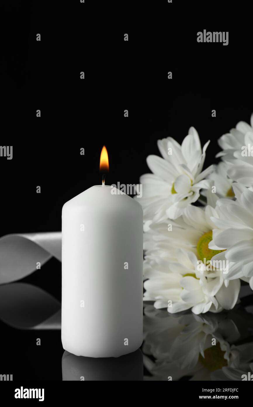 Brennende Kerze, weiße Chrysanthemen und Band auf schwarzer Spiegeloberfläche in Dunkelheit, Nahaufnahme mit Platz für Text. Begräbnissymbole Stockfoto