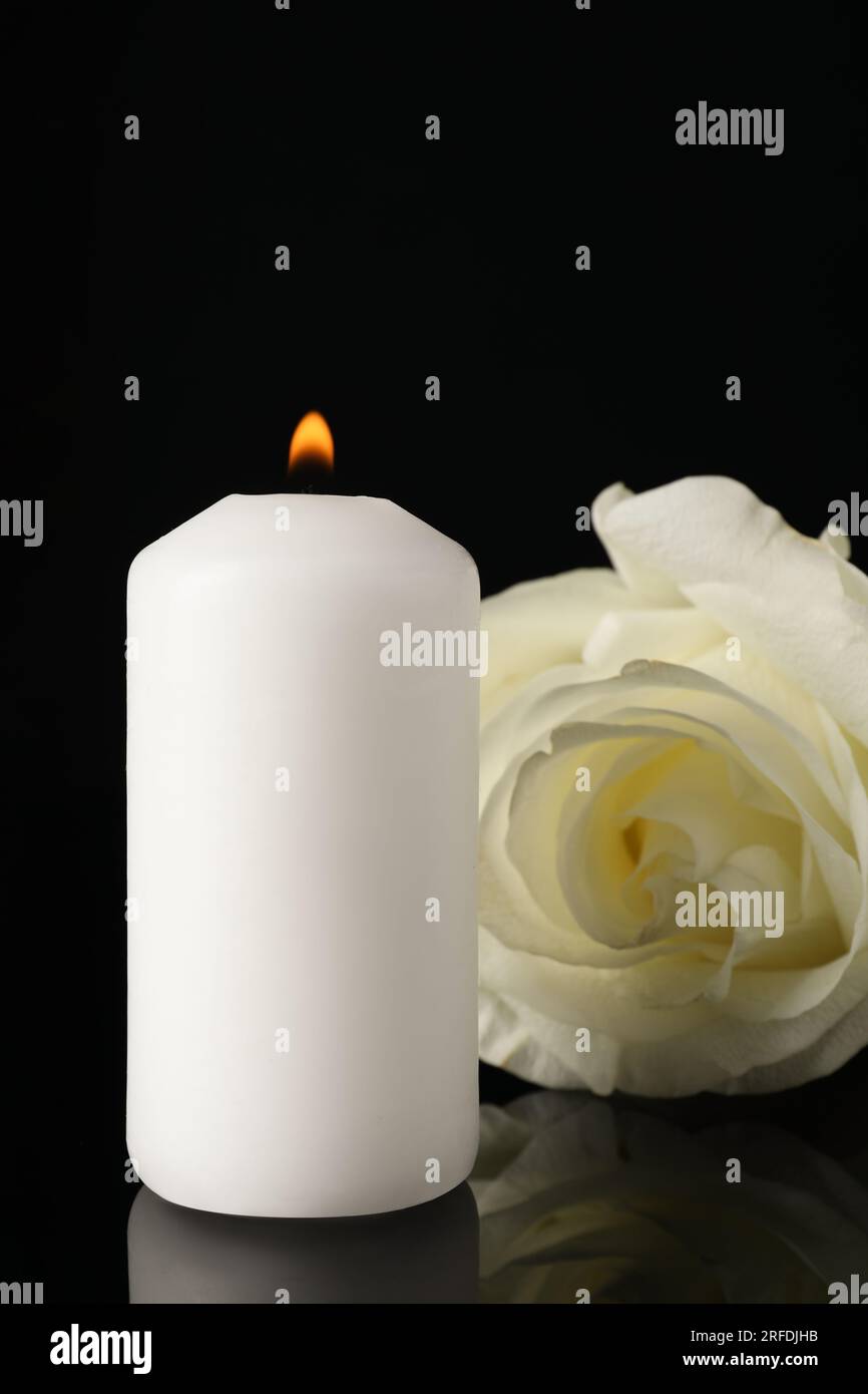 Weiße Rose und brennende Kerze auf schwarzer Spiegeloberfläche in der Dunkelheit, Nahaufnahme. Begräbnissymbole Stockfoto