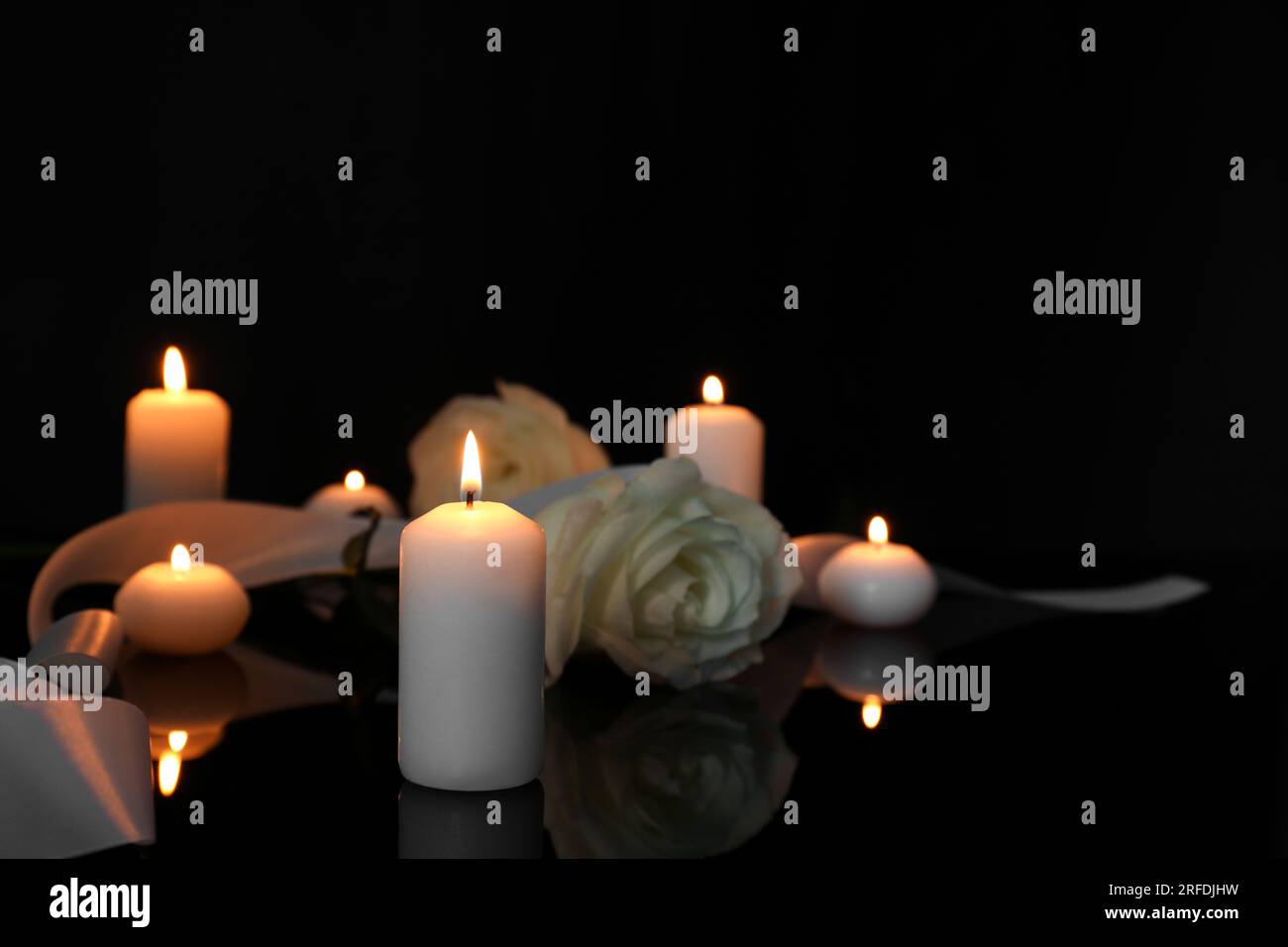 Weiße Rosen und brennende Kerzen auf schwarzer Spiegeloberfläche bei Dunkelheit, Platz für Text. Begräbnissymbole Stockfoto