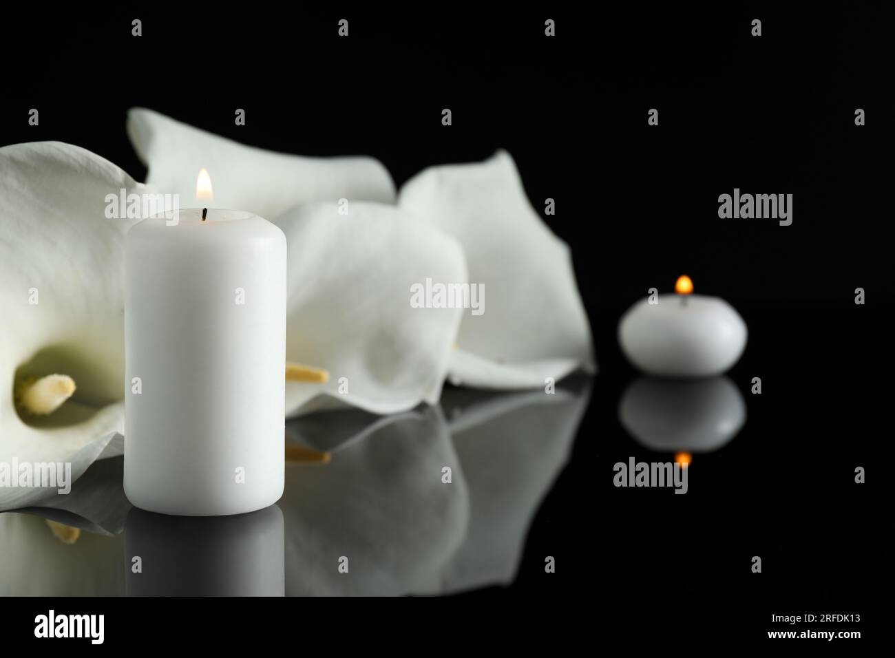 Weiße Calla-Lilien-Blumen und brennende Kerzen auf schwarzer Spiegeloberfläche in Dunkelheit, Nahaufnahme mit Platz für Text. Begräbnissymbole Stockfoto