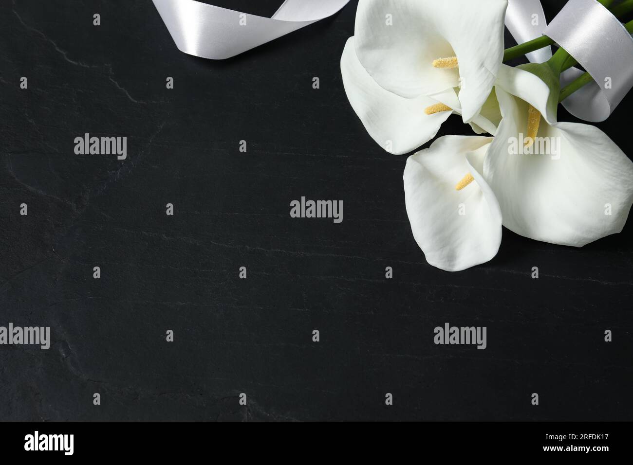 Wunderschöne Calla Lilien Blumen und weißes Band auf schwarzem Tisch, über der Ansicht mit Platz für Text. Begräbnissymbole Stockfoto