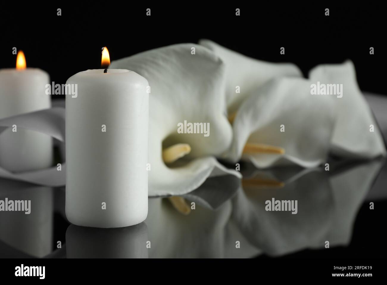 Brennende Kerze und weiße Calla-Lilien-Blüten auf schwarzer Spiegeloberfläche bei Dunkelheit, Nahaufnahme mit Platz für Text. Begräbnissymbole Stockfoto