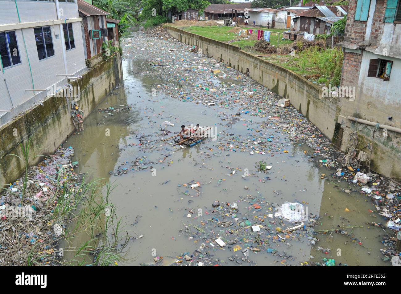 Ein Vater und Sohn rudern auf einem handgefertigten Floß und suchen nach Altkunststoff-Recyclingprodukten aus einem mit Kunststoffabfällen gefüllten Kanal im Kazir Bazar-Viertel von Sylhet. Dieser Kanal ist direkt mit dem Fluss Surma verbunden, der den Fluss Surma verschmutzt und das Flussbett füllt. Das Polyethylen hat sich in riesigen Haufen Schlamm angesammelt, was die Navigation behindert und zu einem Mangel an nutzbarem Wasser aus dem Surma-Fluss von Bangladesch führt. Sylhet, Bangladesch. Stockfoto