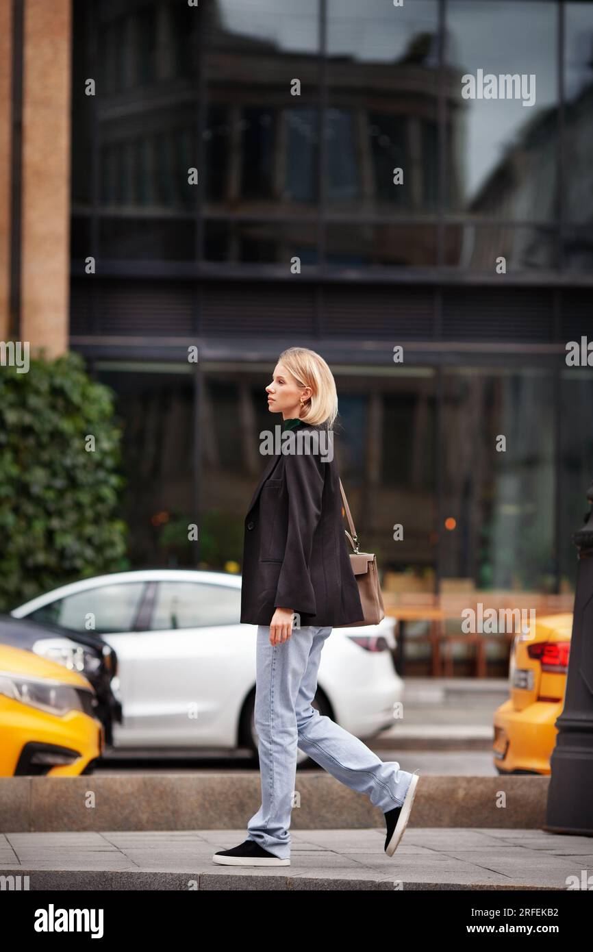 Wunderschöne junge, stilvolle Frau, die vor dem Hintergrund des Gebäudes die Straße entlang läuft, modische Kleiderjeans, schwarzen Blazer, Handtasche und Schnauze trägt Stockfoto