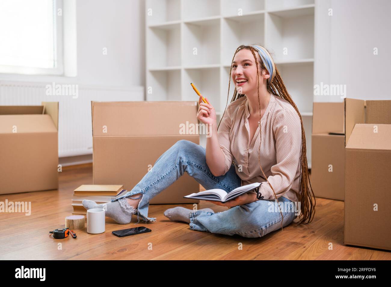 Moderne Ingwerfrau mit Zöpfen, die im Notebook schreiben, während sie in ein neues Zuhause zieht. Stockfoto