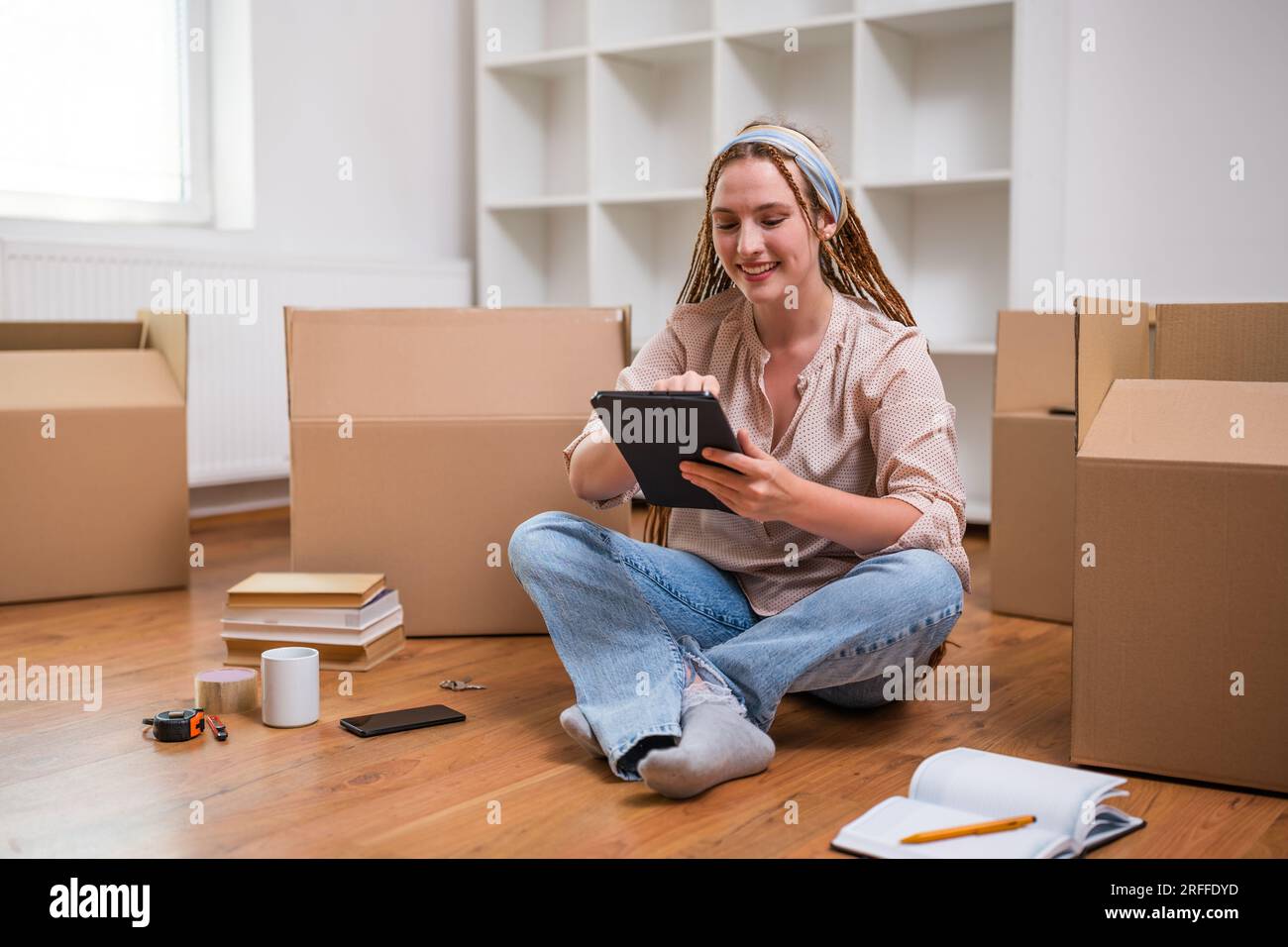 Moderne Ingwerfrau mit Zöpfen auf digitalem Tablet beim Einzug in eine neue Wohnung. Stockfoto