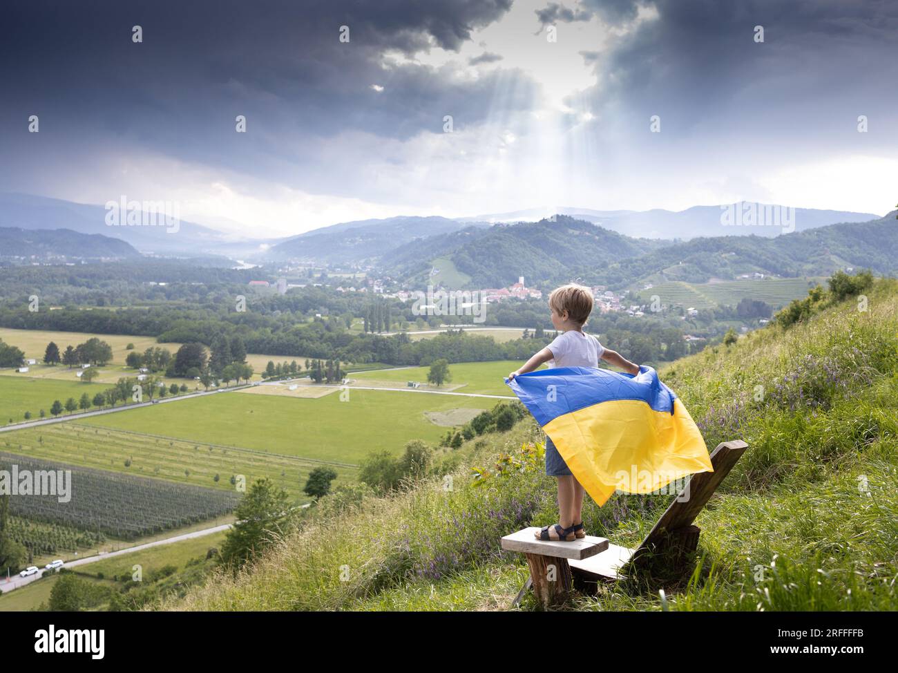Der Junge steht auf dem Berg mit der blauen gelben ukrainischen Flagge, die hinter ihm flattert, vor ihm ist eine wunderschöne Landschaft. Stolz, Ukrainer zu sein. Sei vernünftig Stockfoto