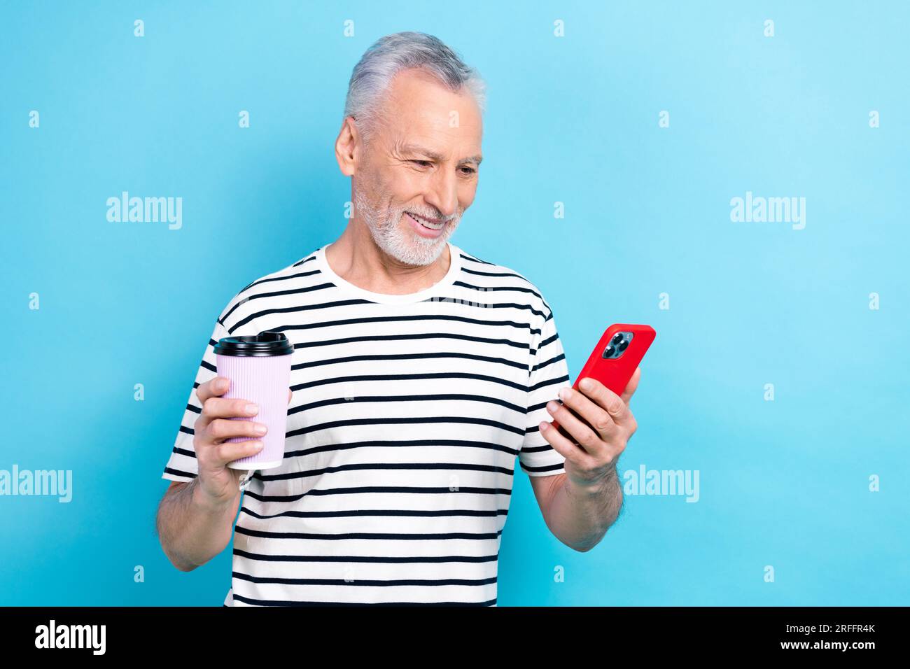 Foto eines lächelnden, fröhlichen Rentners Tragen Sie trendige Kleidung trinken Kaffee Verwenden Sie Telefon iphone Lesen Sie Nachrichten isoliert auf blauem Hintergrund Stockfoto