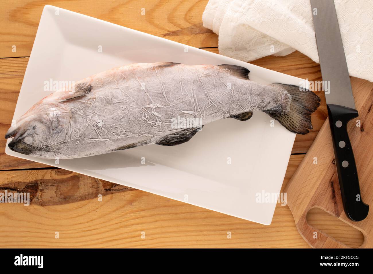 Eine gefrorene Forelle mit weißer Keramikplatte, Messer und Leinenserviette auf einem Holztisch, Makro, Draufsicht. Stockfoto