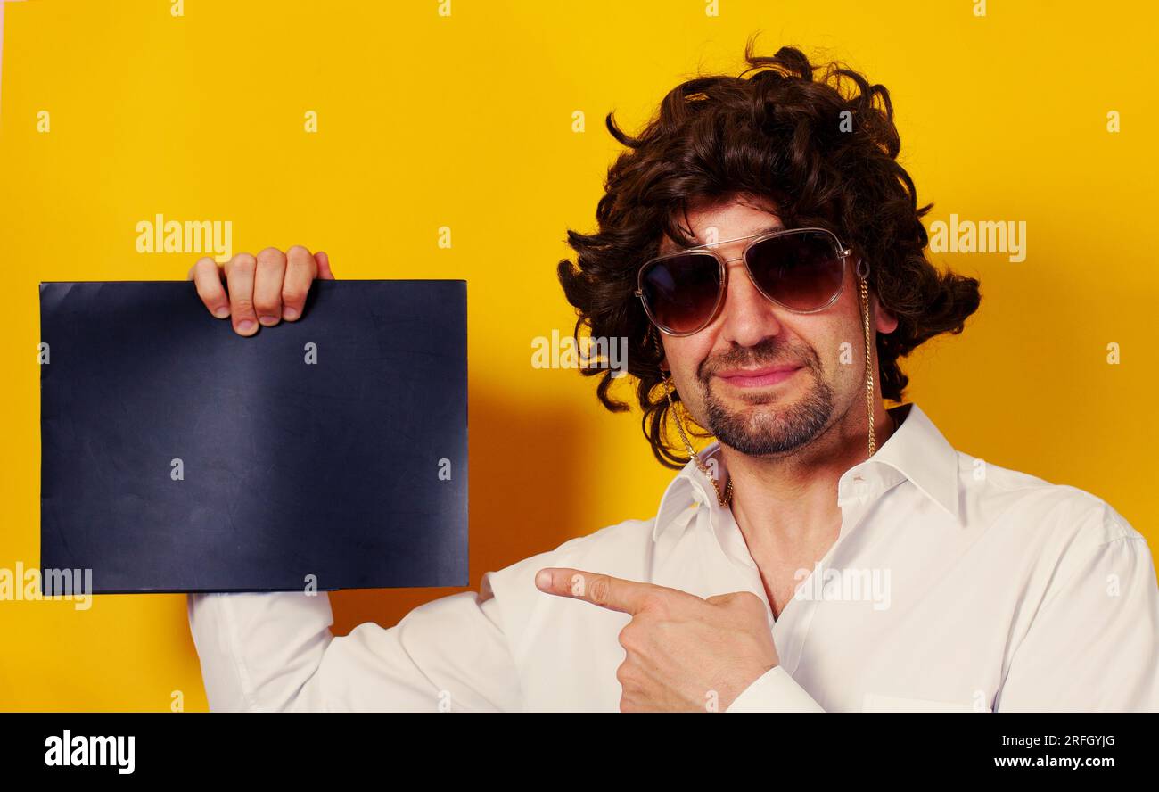 Lockiger, bärtiger junger Mann, der lächelt und eine Sonnenbrille trägt und auf ein schwarzes, leeres Laken auf gelbem Hintergrund zeigt. Platz für Text . Studioaufnahme. Stockfoto