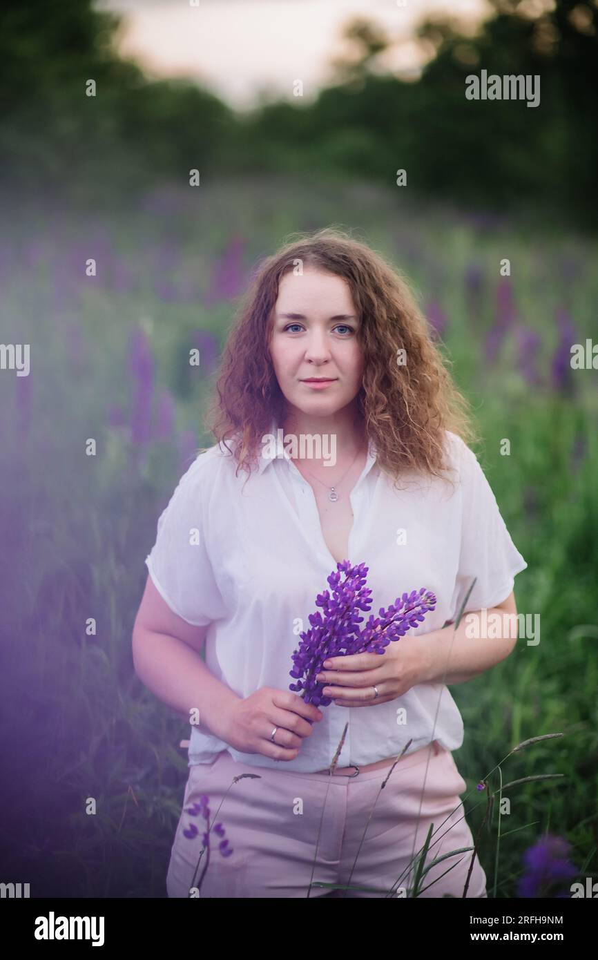 Junge Frau steht in einem weißen Hemd im Feld der lila und rosa Lupinen. Wunderschöne junge Frau mit lockigem Haar draußen auf einer Wiese, Lupinen blühen. Sonnen Stockfoto