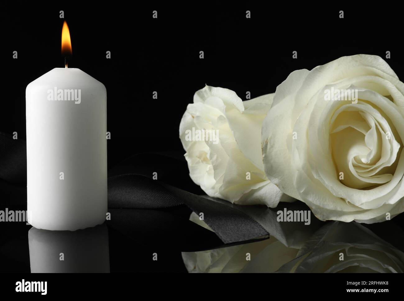 Brennende Kerze, weiße Rosen und Band auf schwarzer Spiegeloberfläche in Dunkelheit, Nahaufnahme. Begräbnissymbole Stockfoto