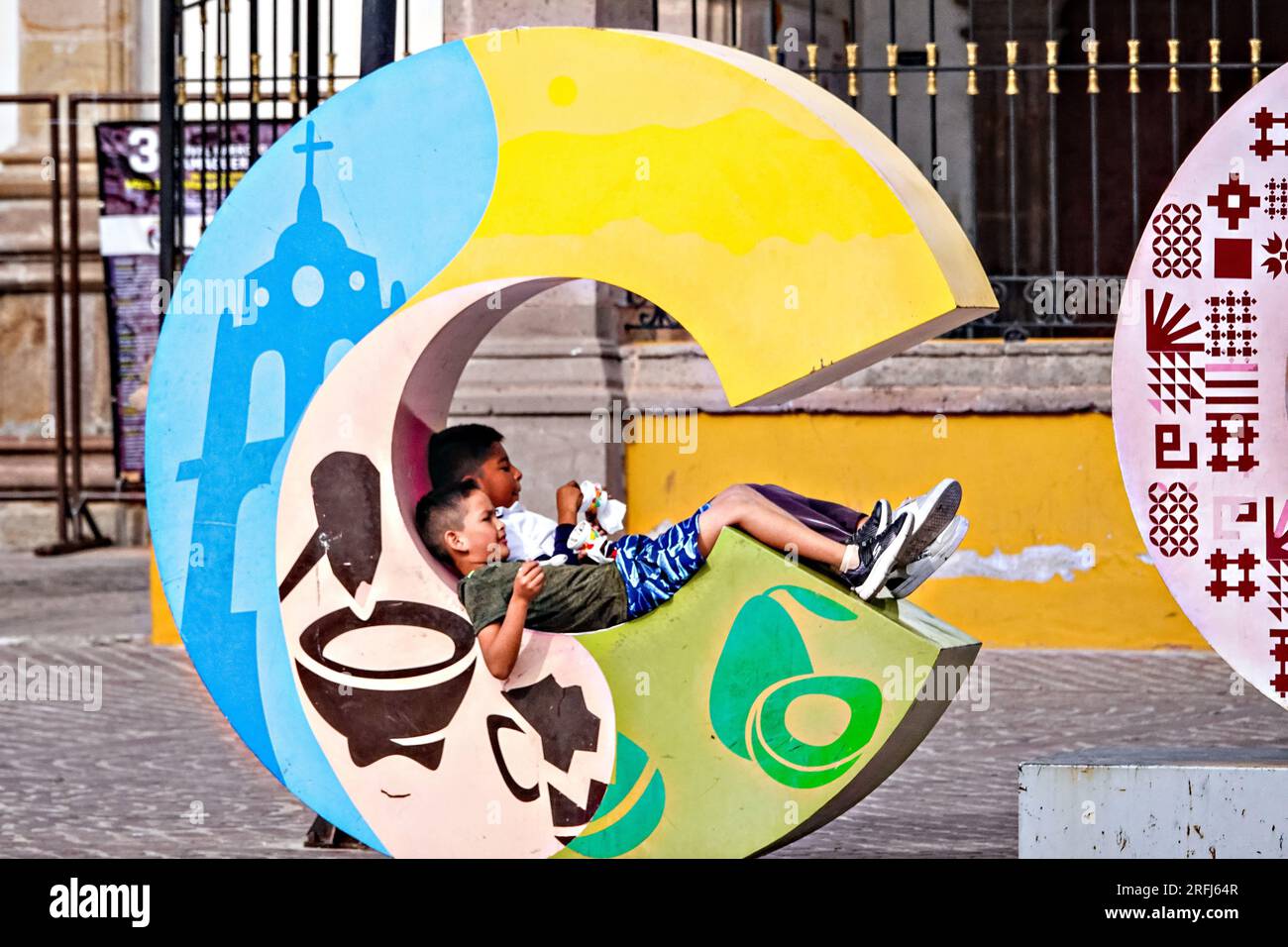 Kleine Kinder entspannen sich auf dem Schild „C“ des Comonfort City am plaza Doctor Mora im historischen Stadtzentrum von Comonfort, Staat Guanajuato, Mexiko. Stockfoto