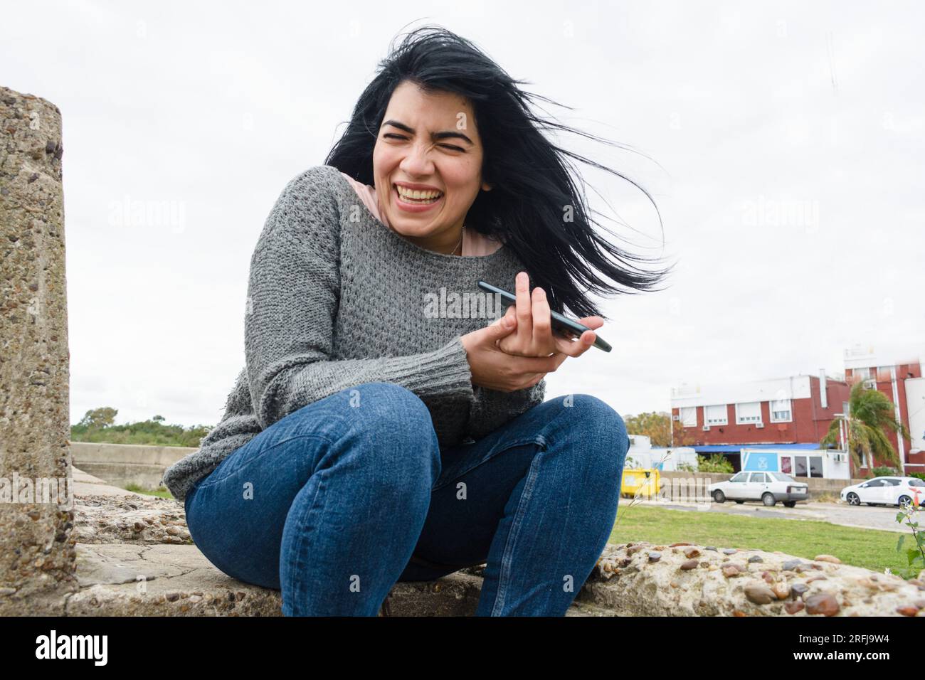 Eine junge latinatin, die glücklich am Telefon lacht, in sozialen Netzwerken surft, Nachrichten und Videos ansieht, draußen sitzt, sich ausruht, sehr unterhaltsam Stockfoto