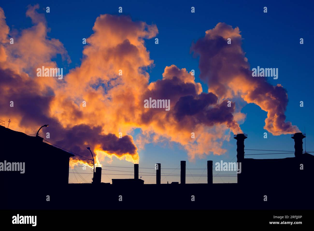 Fabrikschornsteine rauchen giftige Substanzen über der Silhouette von Industriegebäuden über dem wunderschönen blauen Himmel bei Sonnenuntergang oder Sonnenaufgang Stockfoto