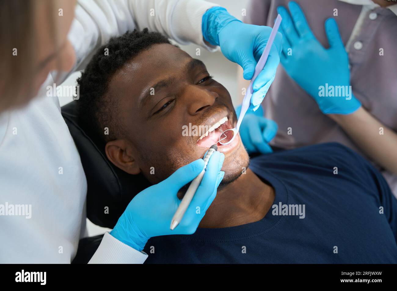 Afroamerikanischer männlicher Mandant liegt auf dem Zahnstuhl während eine Frau Stomatologin die Gesundheit seiner Mundhöhle überprüft mit Mundspiegel und Sichelsonde Stockfoto