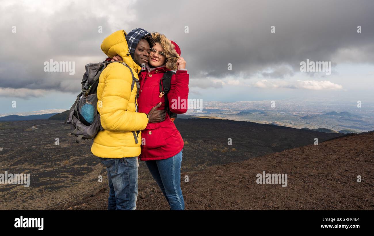 Ein multiethnisches junges Paar, ein afrikanischer Mann und eine weiße Frau, genießen Sie einen Berggipfel mit Panoramablick. Herunterziehende Jacken und technischer Rucksack Stockfoto