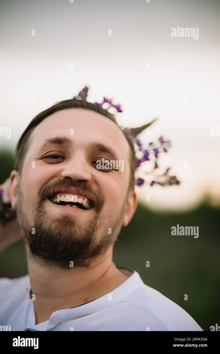 Junger lächelnder Mann mit einem Strauß Lupinen wie eine Krone. Sonnenuntergang oder Sonnenaufgang, helles Abendlicht. Stockfoto