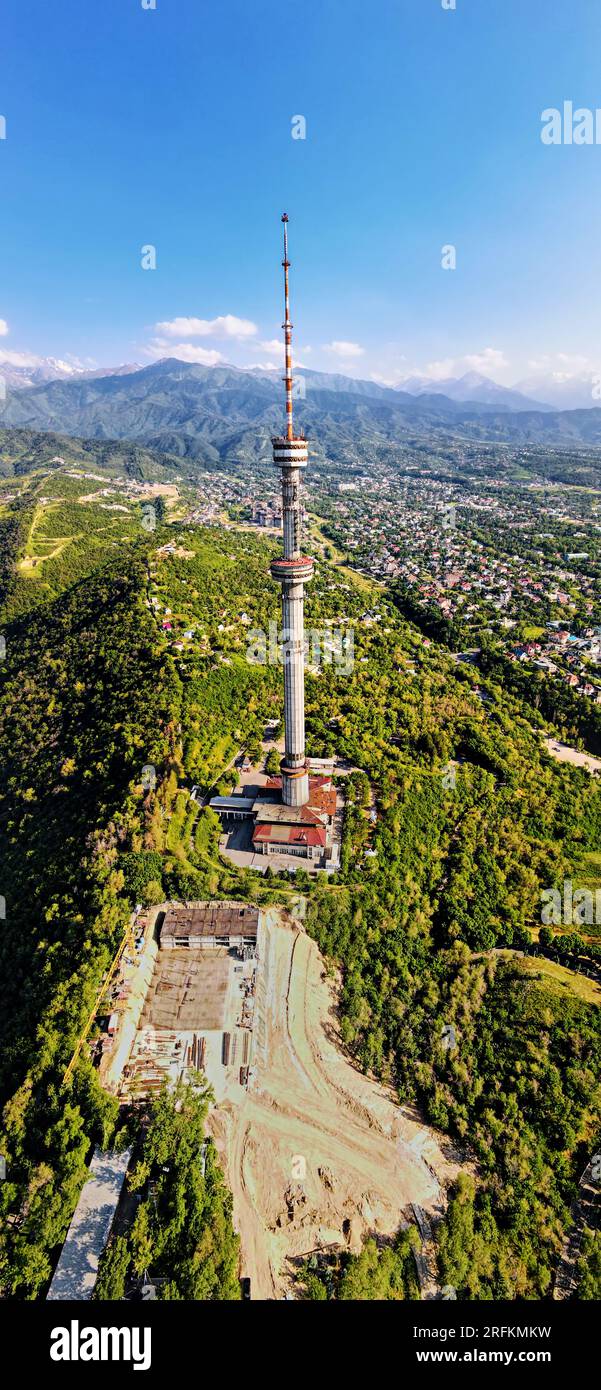 Vertikale Luftdrohnenaufnahme des Hochfernsehturms der Stadt Symbol Almaty und Parken auf dem Koktobe-Hügel vor den Schneebirgen Kasachstans Stockfoto