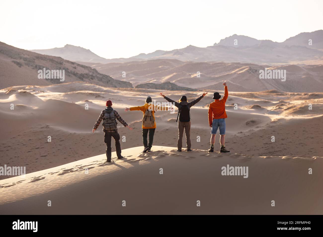 Vier glückliche Touristen mit Rucksäcken stehen auf Sanddünen mit offenen Armen und blicken auf den Sonnenuntergang Stockfoto