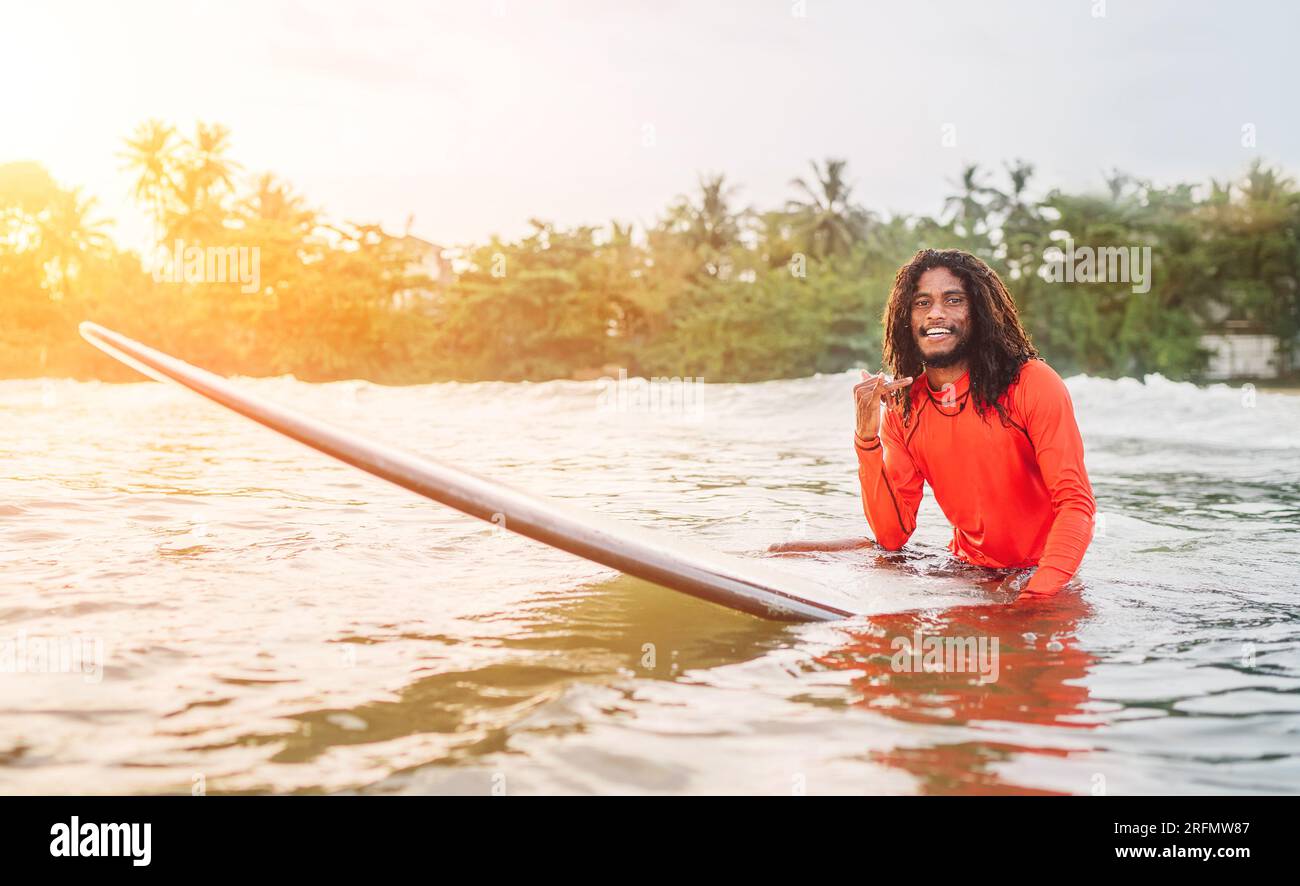 Schwarzer langhaariger Teenager-Mann, der shaka-Schild auf einem langen Surfbrett zeigt und auf Wellenreiten wartet, mit Palmen gesäumten Sonnenstrahlen. Extreme Wasserspor Stockfoto