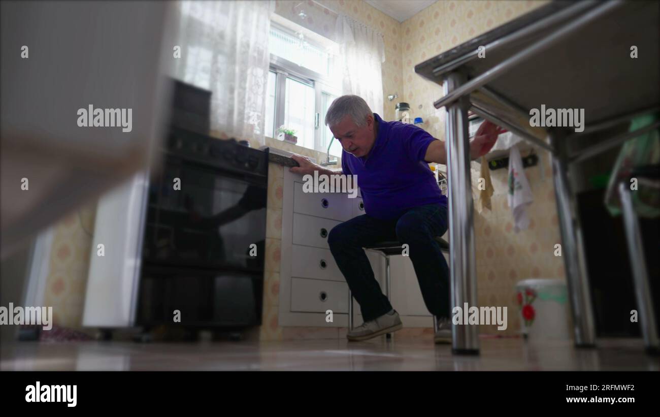 Älterer Mann mit Herzinfarkt, der auf den Boden fällt und einen medizinischen Notfall zu Hause benötigt, durch Küchenspüle. Kranker Seniorenarzt mit Herz-Kreislauf-Erkrankungen Stockfoto