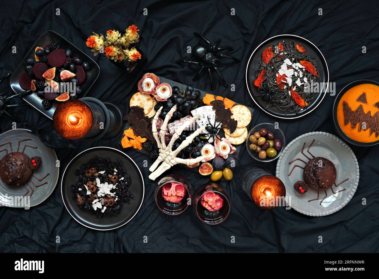 Halloween-Dinner-Party-Tischszene auf dunklem schwarzen Hintergrund. Ansicht von oben nach unten. Gruselige Wurstplatte, schwarzes Risotto und Pasta, Spinnenkuchen, Jack o Stockfoto