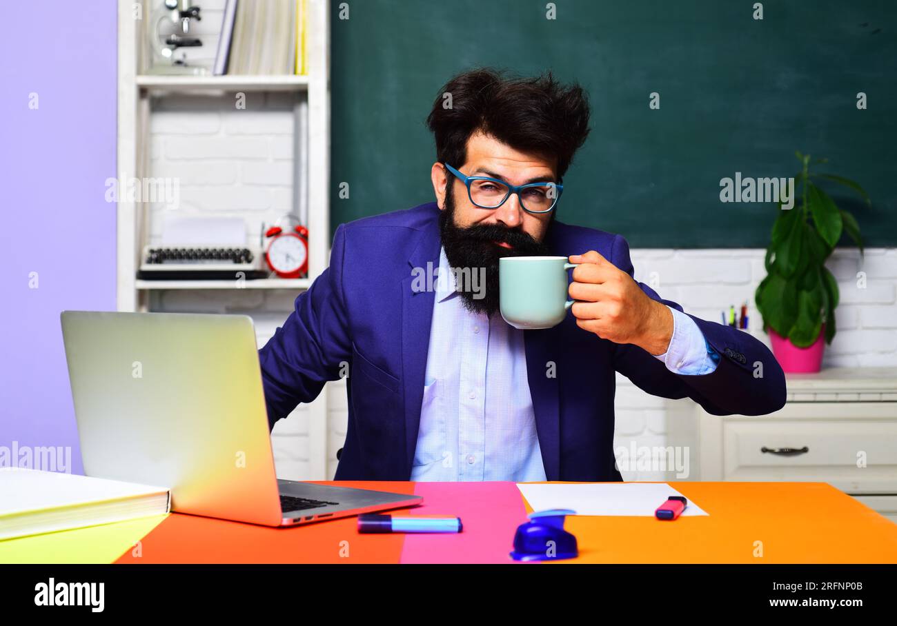 Schläfriger männlicher Student, der mit einem Laptop arbeitet und Kaffee im Klassenzimmer trinkt. Lehrer oder Universitätsprofessor am Arbeitsplatz, der eine Tasse Tee trinkt. Ich Bin Müde Stockfoto
